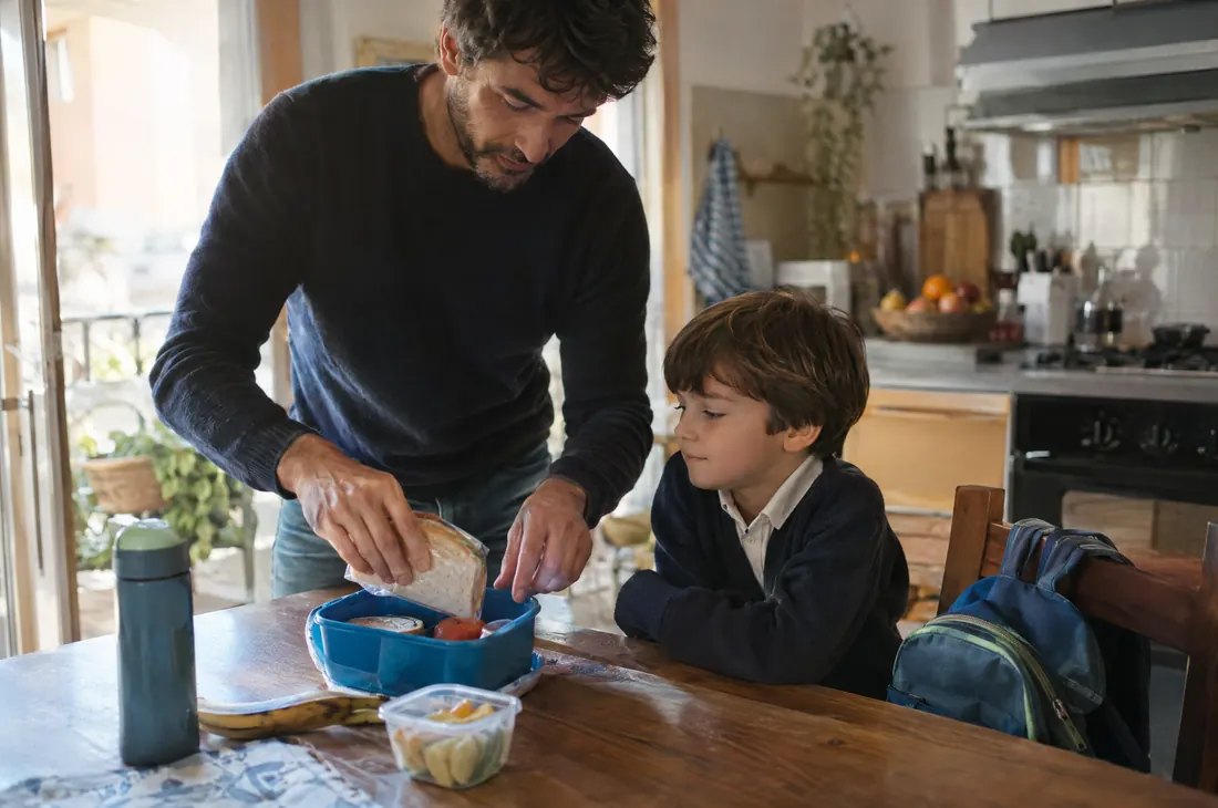 Un padre prepara la merenda per la scuola al figlio, con lo zaino appoggiato alla sedia
